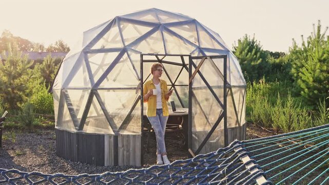 Caucasian woman stand in a unique geodesic dome gazebo, hold a mobile phone and a cup. Relaxed outdoor setting with natural light, ideal for leisure, lifestyle, or technology promote