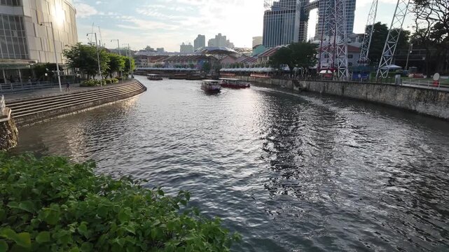 Scenic video capturing a river cruise boat gliding along the Singapore River near Clarke Quay, surrounded by waterfront architecture, modern skyscrapers, reflections on calm water, and warm evening li
