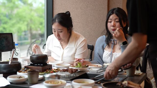 Three women enjoy a shared meal together. They savor food with chopsticks and small pots. One woman smiles, another looks focused. They sit near a large window with green foliage