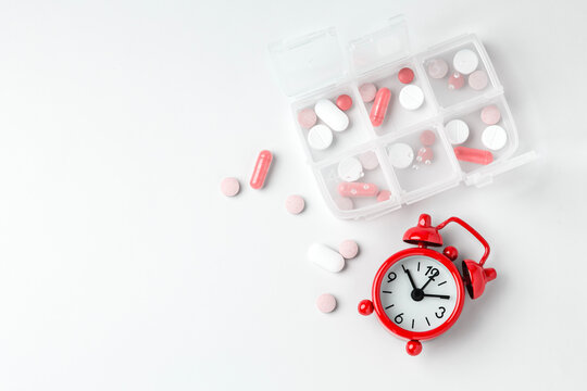 Pill organizer box with vitamins and supplements and red alarm clock on white background, top view