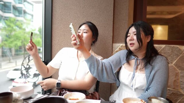 Two women enjoy a meal together. One holds phone, capturing selfie. Other laughs softly, looking at screen. They sit near window, sipping tea. Cozy restaurant, warm, inviting