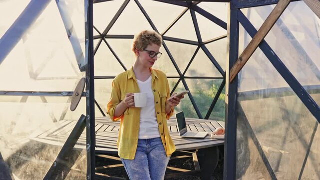 A smiling Caucasian woman with short blonde hair and glasses holds a mug while talking on her phone inside gazebo greenhouse