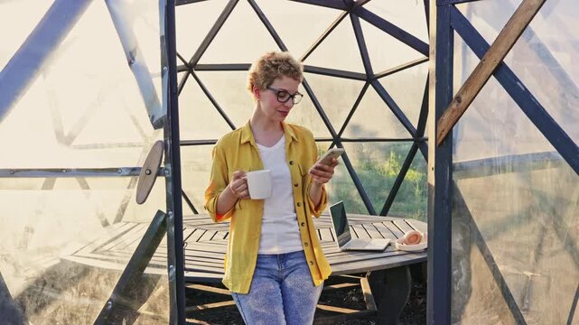 A Caucasian woman with short blonde hair and glasses holds a mug and smartphone while standing inside a gazebo greenhouse