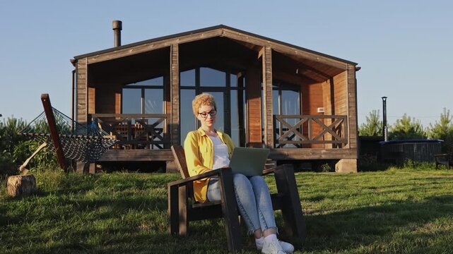 A caucasian woman with short blonde hair and glasses sits in a chair outdoors with a laptop, enjoying a sunny day near a wooden cabin