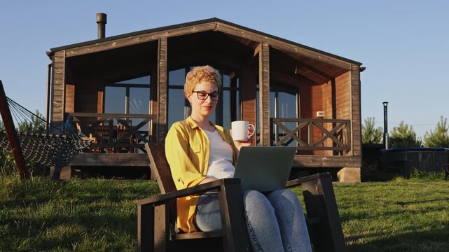 A caucasian woman enjoys a cup of coffee while working on her laptop in a chair outside her wooden cabin