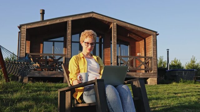 A happy woman with short blonde hair and glasses sits in a chair outside a wooden cabin, raising her fist in triumph while holding a mug and looking at her laptop
