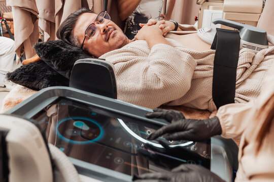 Middle-aged man relaxing during an Emsculpt HIFEM body contouring session while an aesthetic practitioner adjusts treatment settings on the device in a modern medical spa.