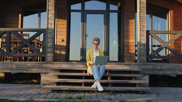 A young caucasian woman with curly hair sits on wooden steps in front of a modern wooden house, typing on her laptop computer