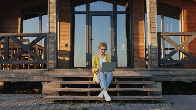 A joyful caucasian woman with curly hair sits on wooden steps outside a cabin, enthusiastically celebrating a success on her laptop