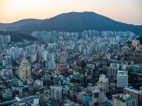 Aerial view of the dense urban cityscape of Busan featuring high-rise apartment buildings and a mountain backdrop under a soft sunset sky in Busan, Busan, South Korea.