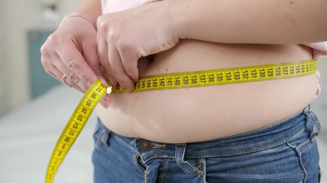Close-up of a person measuring waist circumference with a measuring tape to monitor body fat, obesity, weight loss, fitness progress, and healthy lifestyle goals.