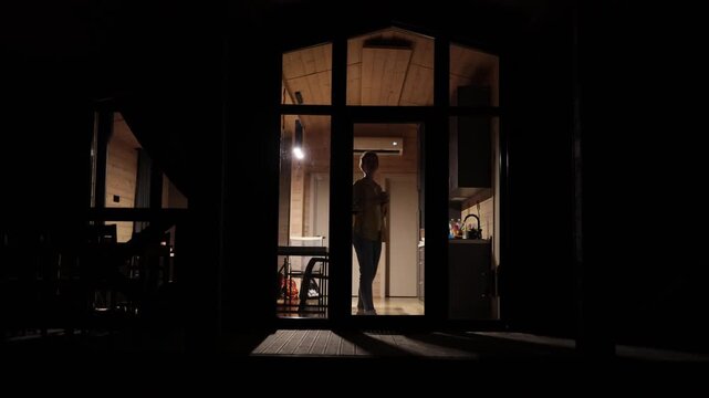 A silhouette of a caucasian woman holding a mug stands in a warmly lit kitchen at night, looking out into the darkness