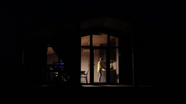 A young woman stands in a brightly lit kitchen at night, illuminated by interior lights while working on a counter
