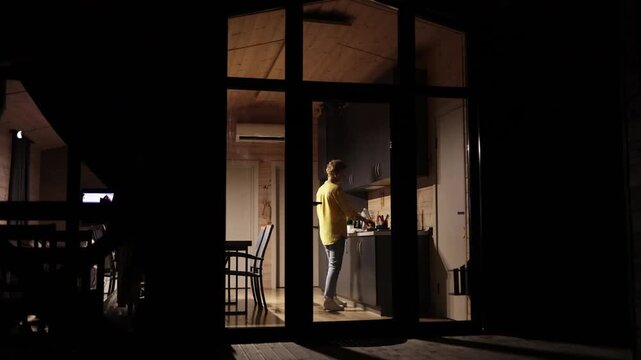 A young woman stands in a modern kitchen at night, illuminated by interior lights, preparing food