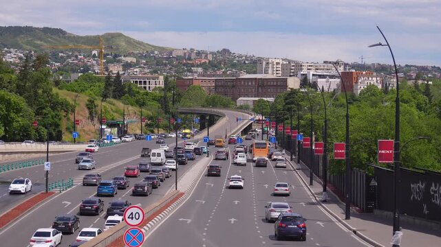 Aerial view of cars and vans driving on a busy highway along the embankment of the muddy mtkvari river in tbilisi, the capital of georgia, surrounded by lush green trees and city buildings