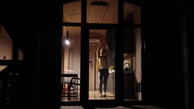 A young caucasian woman joyfully dances with her arms raised inside a contemporary wooden cabin kitchen at night