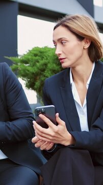 Sad disinterested business man and woman looking disappointedly at uninteresting information on their smartphones while sitting outside on a modern office background. Disappointed business people in