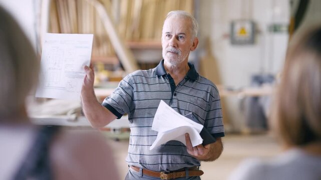 Elderly foreman holding paper blueprints explaining project details to colleagues. Experienced mentor sharing knowledge with workers in a carpentry