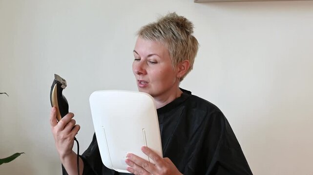 A woman shaves her hair with a trimmer and holds mirror. Balding hair loss due to chemotherapy. Baldness in women during cancer treatment. Toxic effects of chemotherapy drugs on hair.