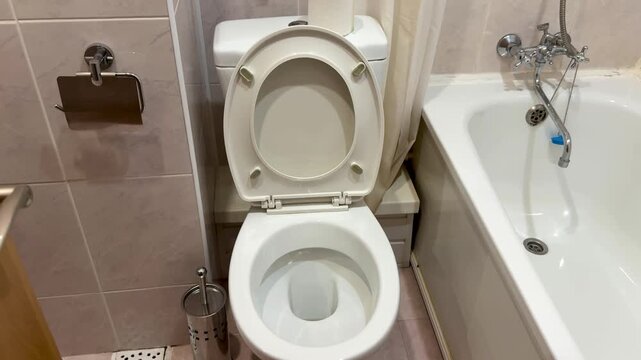 Close-up white toilet and bathtub, economy hotel bathroom with beige tiles, chrome faucet and shower hose, curtain tucked to side