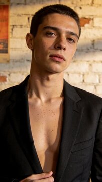 Vertical video: Posing on cue, man gripping black blazer, tilting head for shoot against brick wall