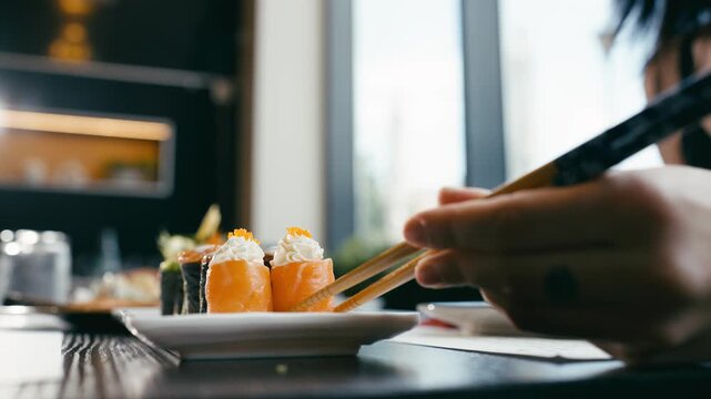 Close up of a person using chopsticks to pick up a salmon sushi roll with cream cheese from a plate in a modern Japanese restaurant