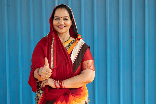 Portrait of a smiling Indian woman showing a thumbs-up sign against a blue background. Positive expression representing success, motivation, women empowerment, social development, rural leadership.