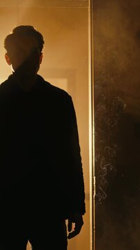 Vertical video: Standing man in dark jacket moving hand at doorway, warm light creating smoky halo