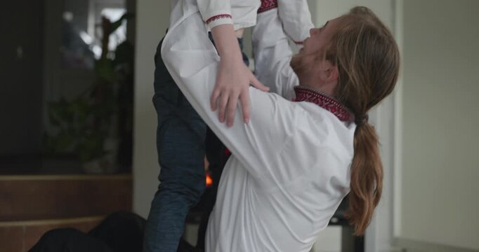 Man Hugs Embraces Kid Boy Have Fun Together on Floor Near Fireplace. Happy Family in Ukrainian Traditional Attire. 4K 10-bit