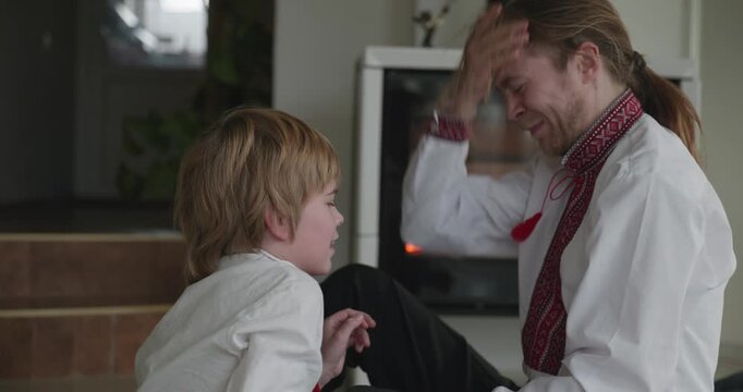 Man Hugs Embraces Kid Boy Have Fun Together on Floor Near Fireplace. Happy Family in Ukrainian Traditional Attire. 4K 10-bit