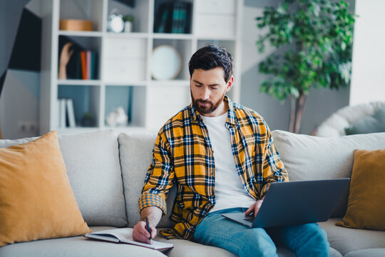 Young man working from home on a couch with laptop and notebook, writing notes and concentrating while studying or freelancing in a bright casual living room
