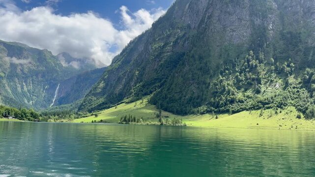 Panoramic, scenic view of Konigssee Lake. Rothbach waterfall in the background. Salet stop where route to Obersee starts. View from a moving boat. Chrystal clear water. Konigssee, Bavaria, Germany
