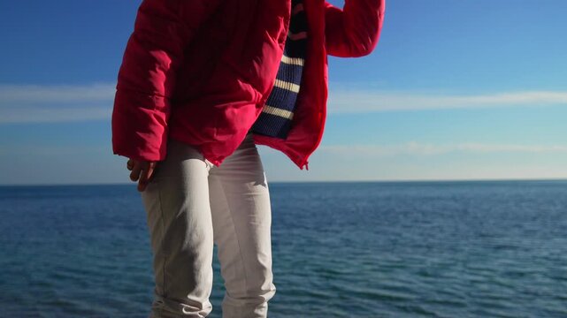 Woman throwing stone into the calm blue sea water at the beach while wearing a bright pink puffer jacket on a sunny day