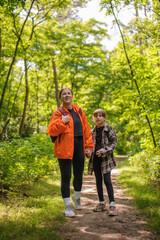 Fototapeta premium Smiling mother and daughter with backpacks holding hands while standing on trail in forest 
