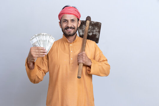 Portrait of smiling young farmer showing indian rupees currency wearing kurta pajama with gamcha gesturing  and holding spade looking athe camera while standing against white background