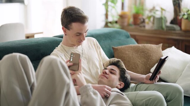 Two young men relaxing on a couch. One man is lying on his boyfriend's lap using a smartphone, while the other is holding a tablet