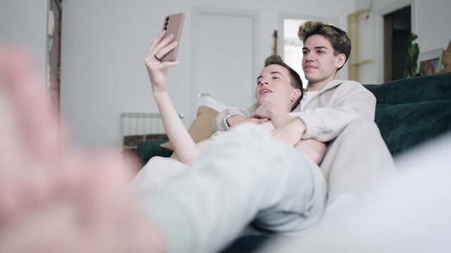 Young gay couple cuddling and having fun on the sofa at home. One man holds a smartphone and talks during a video call with friends