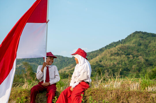 Two Indonesian school children sitting in a sunny field holding the national flag of Indonesia, celebrating patriotism, freedom. Hari Kemerdekaan, Merdeka, and Dirgahayu Indonesia on 17 Agustus