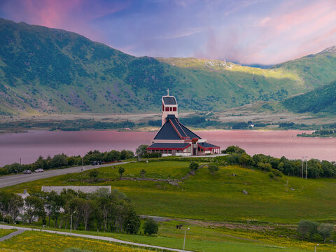 Modern white church with steep roof and square bell tower stands on a grassy hill by a calm fjord in Lofoten, Norway, evening light with pink water reflections.