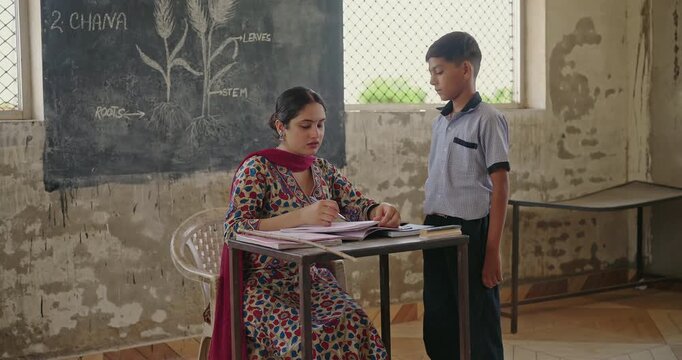 Indian young adult female sitting on chair do work check study book talk speak at class room rural area place. Strict Tutor look angry hold stick hitting teen boy child hand for project notes mistake