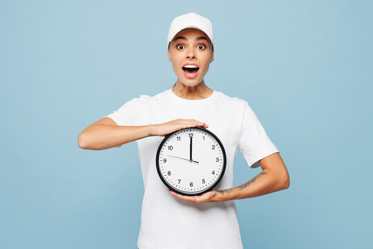 Professional shocked surprised delivery girl employee woman wear white cap t-shirt uniform workwear work as dealer courier hold in hand clock isolated on plain pastel blue background. Service concept.