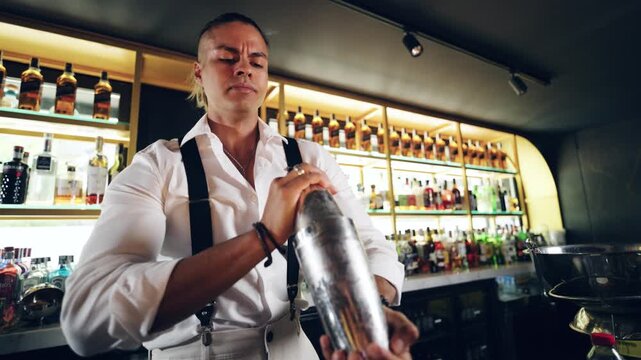 Focused barman in a white shirt and suspenders vigorously shaking a stainless steel cocktail shaker while preparing a drink at a bar counter