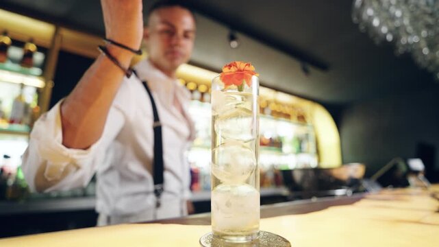 Professional mixologist carefully placing an edible flower garnish on a tall glass filled with ice and a clear beverage at a modern bar