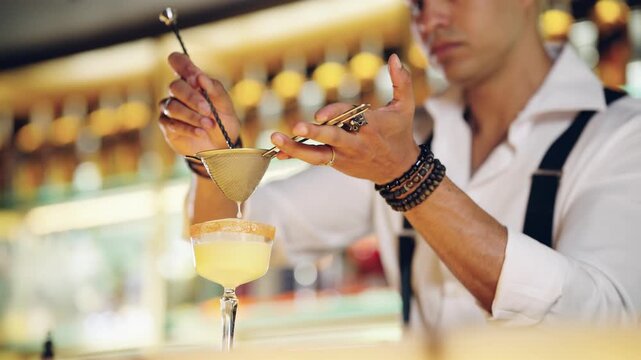 Stylish male bartender carefully preparing a yellow cocktail, pouring the liquid through a fine strainer into an elegant glass at a bar