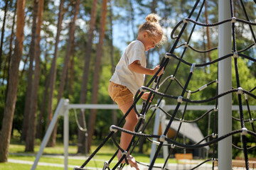 A young child ascends a rope climbing frame at a playground in the park. The sun shines down as trees provide background greenery © sementsova321