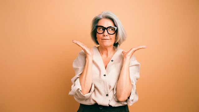 Senior woman in eyeglasses shrugging, looking to the side with a puzzled facial expression on a plain background