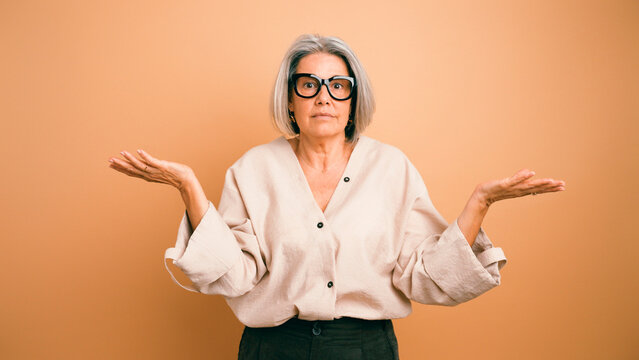 Senior woman with gray hair and glasses shrugging, expressing uncertainty, doubt, and confusion against a peach background