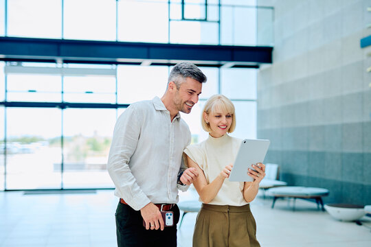Two smiling businesspeople, a man and a woman, collaborating and discussing work using a digital tablet in a bright office lobby