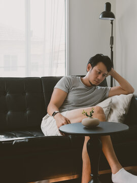 Young Thai man leaning forward tiredly on table from black sofa in relaxed apartment in a quiet minimalist Thai apartment with natural daylight