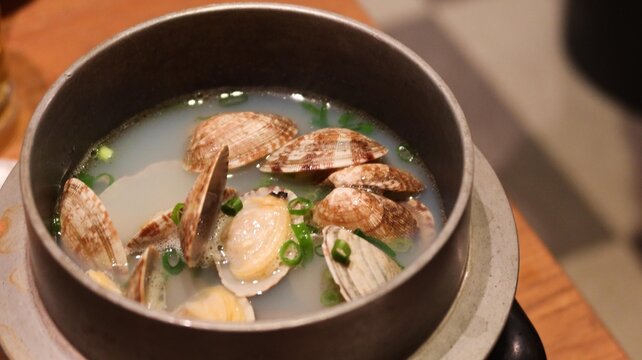 close up of Sake Steamed Clams in a pot served in a traditional metal pot, featuring fresh clams with open shells in a clear, savory broth, garnished with chopped green onions with copy space